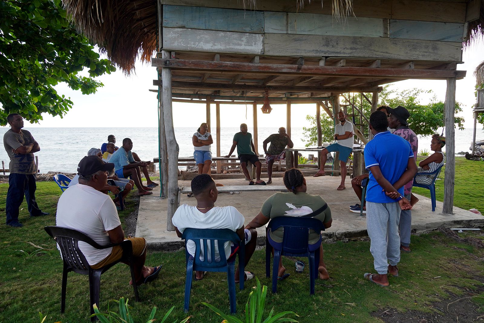 Reunión de comunidad bajo enramada frente al mar, vecinos sentados en círculo